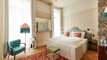 Une chambre avec un grand lit, un fauteuil vert, une petite table avec des bouteilles et un tapis à motifs.