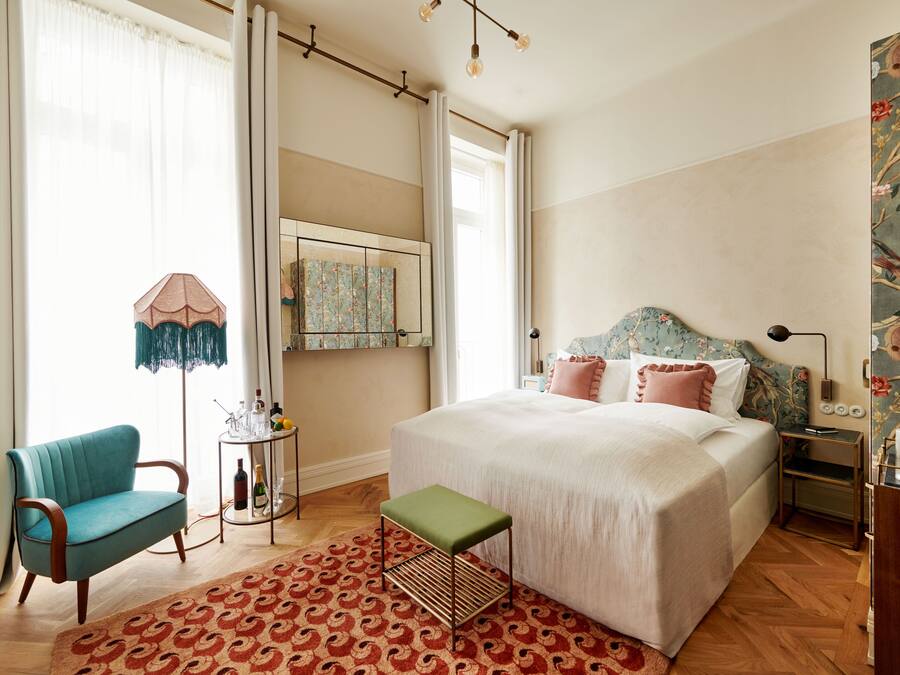Une chambre avec un grand lit, un fauteuil vert, une petite table avec des bouteilles et un tapis à motifs.