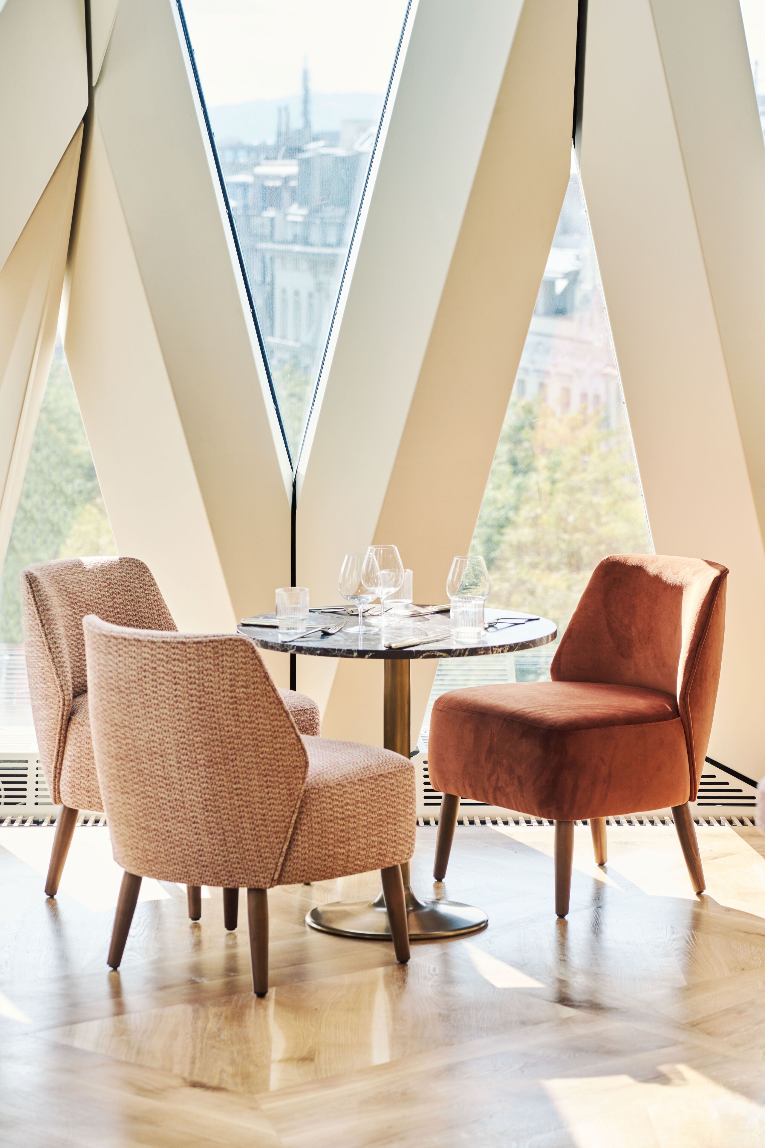 A dining area with a round table, two chairs, and a view of a cityscape through large windows.