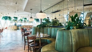 Ein modernes Restaurant mit grünen Polstersesseln, Holztischen, hängenden Pflanzen und Pendelleuchten.