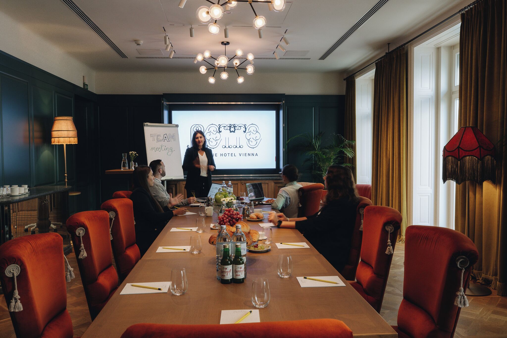 A conference room with a long table, red upholstered chairs, and a projector screen displaying a logo.