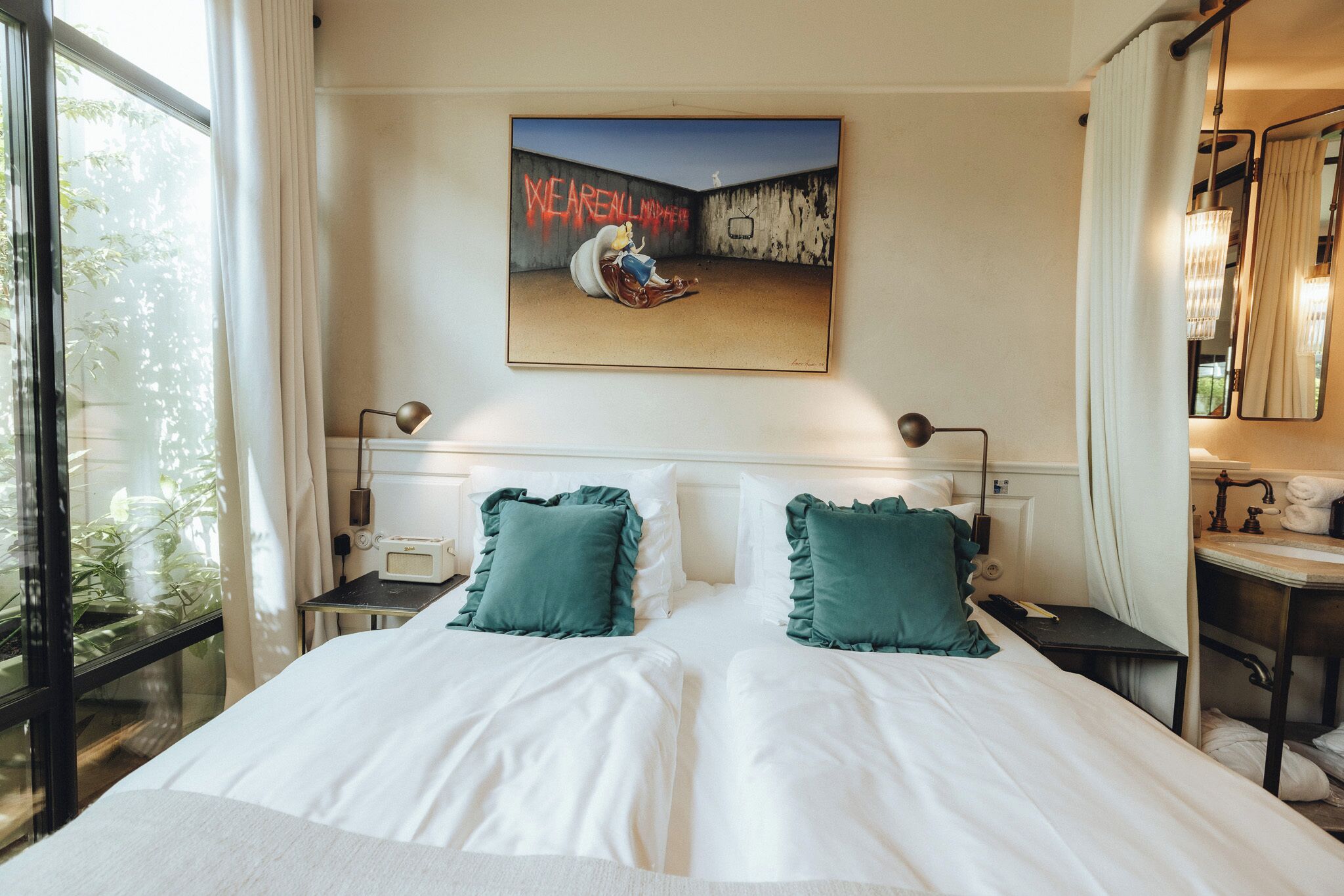 A hotel room with a large bed, two bedside tables, a nightstand, a window with curtains, a painting on the wall, and a bathroom with a mirror and sink.