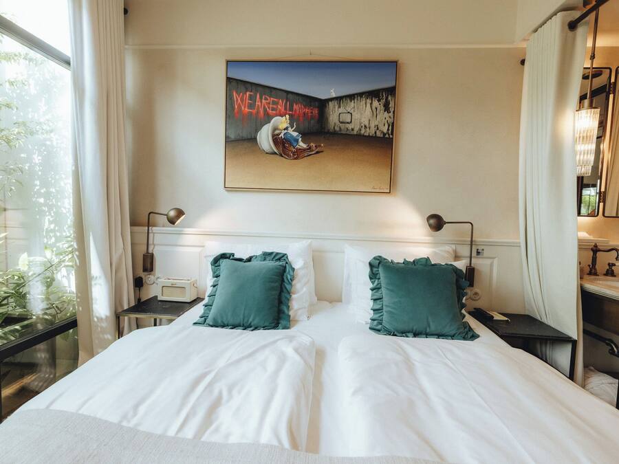 A hotel room with a large bed, two bedside tables, a nightstand, a window with curtains, a painting on the wall, and a bathroom with a mirror and sink.