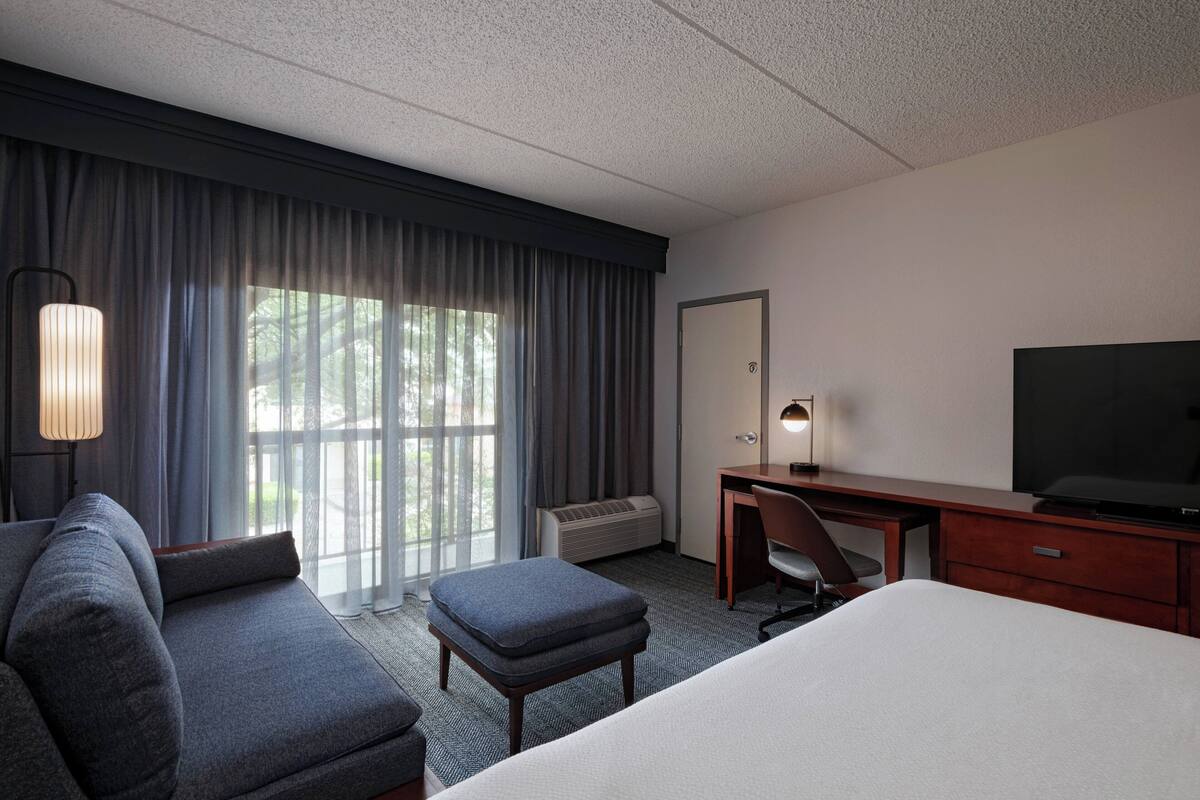A hotel room with a large bed, a sofa, an ottoman, a desk with a chair, a television, and a window with curtains.