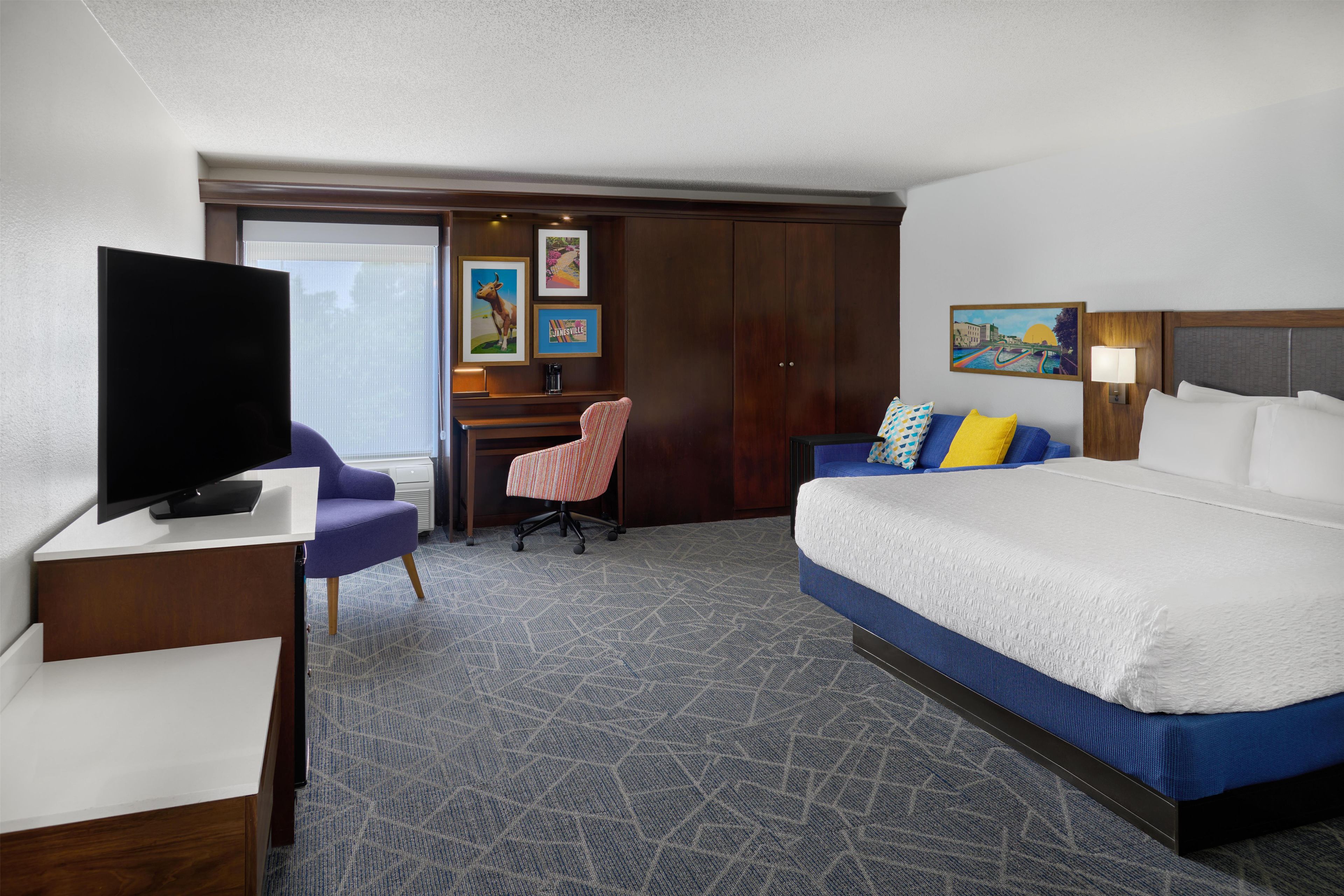 Habitación de hotel con cama, escritorio, televisor, una silla y obras de arte en las paredes.