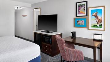 Habitación de hotel con cama, escritorio, silla, televisor de pantalla plana, microondas y cuadros en la pared.