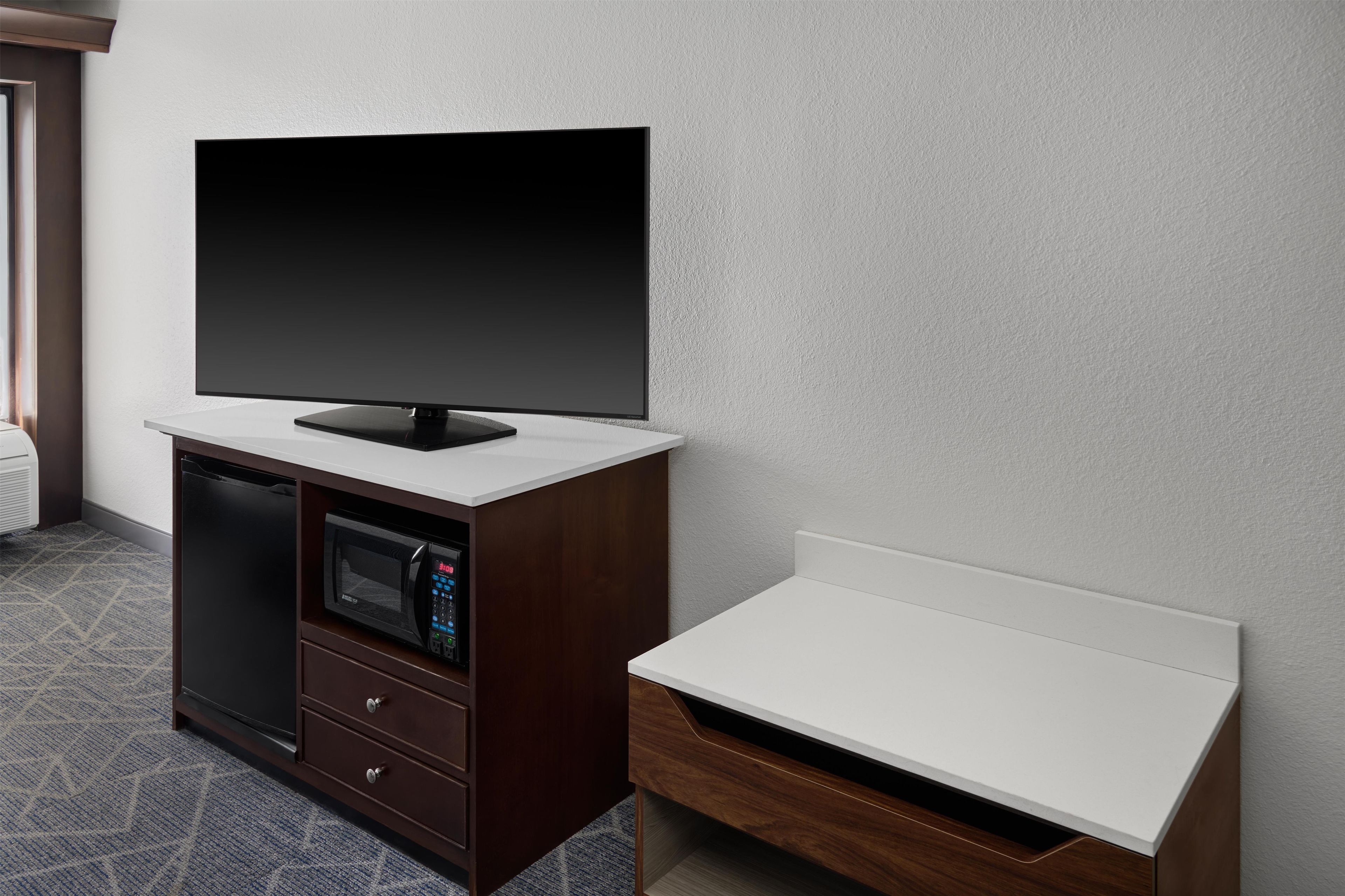A room with a flat-screen TV on a wooden cabinet, a microwave, and a white bedside table.
