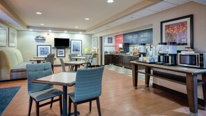 A hotel dining area with tables, chairs, a flat-screen TV, a kitchen counter, and a coffee station. - Hampton Inn Janesville (Janesville)