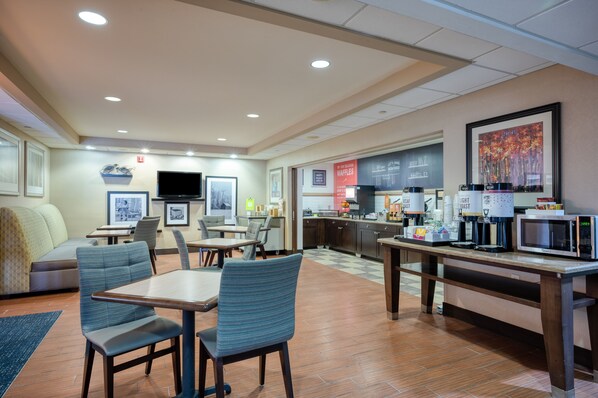 A hotel dining area with tables, chairs, a flat-screen TV, a kitchen counter, and a coffee station. - Hampton Inn Janesville (Janesville)