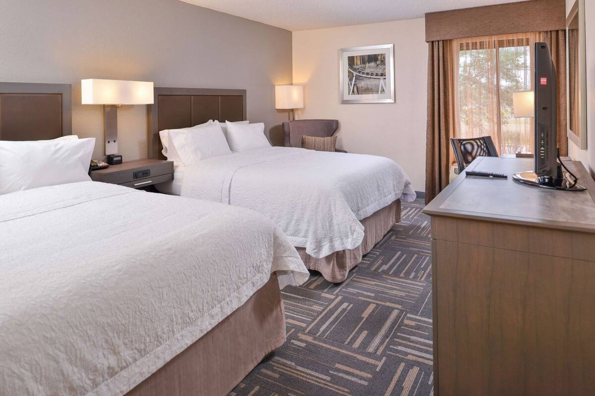 A hotel room with two beds, a desk, a chair, a TV, and a window with curtains.