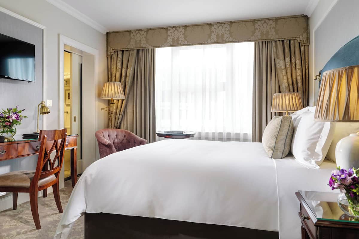 Habitación de hotel con una cama grande, un escritorio con silla, un televisor montado en la pared y una ventana con cortinas.