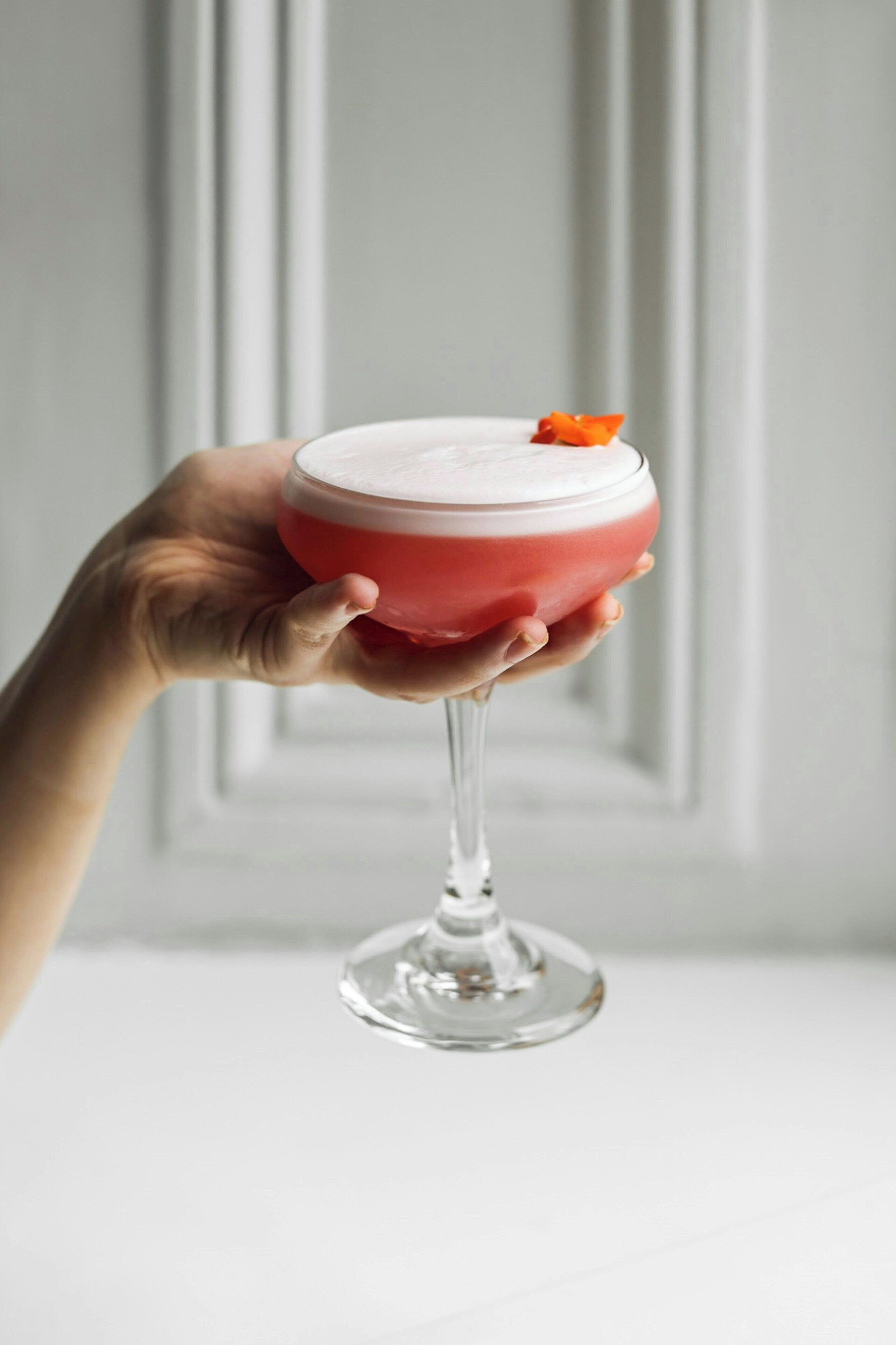 Eine Hand hält ein Cocktailglas mit einem rosafarbenen Getränk und einer weißen Schaumkrone.