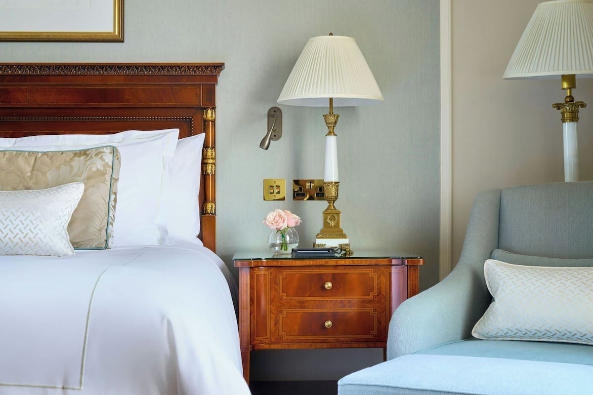 Habitación de hotel con cabecera de madera, mesita de noche con lámpara y zona de estar con sillón acolchado.