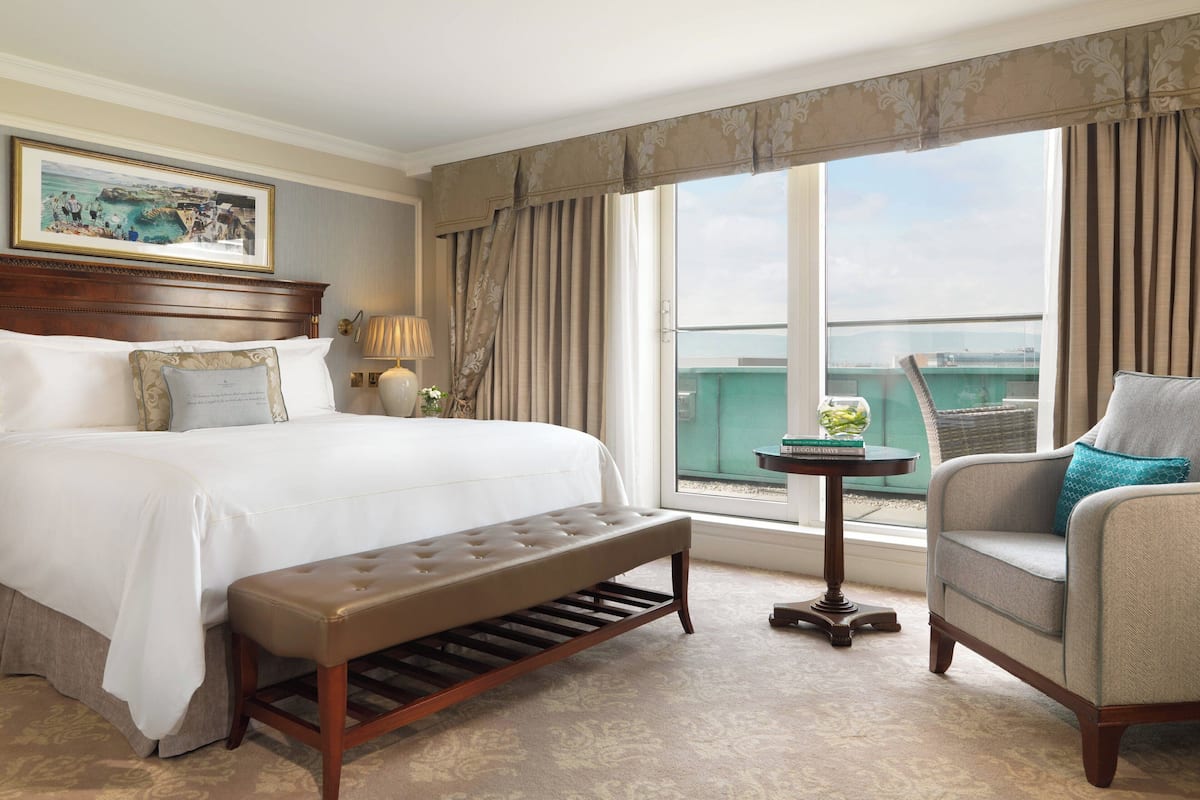 Una habitación de hotel con una cama grande, un banco, una silla, una mesita y vistas a la ciudad a través de la ventana.