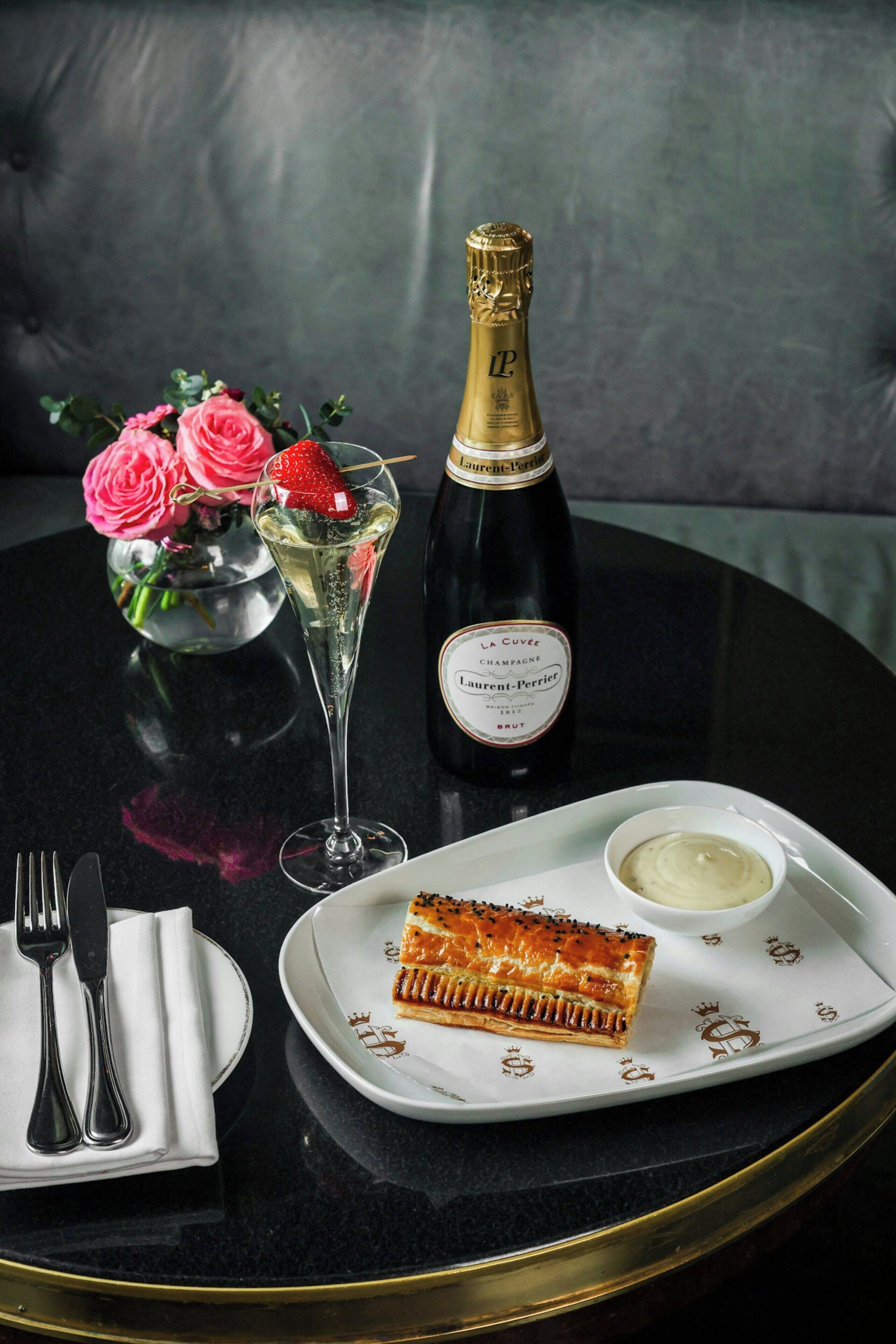 A table with a bottle of champagne, a glass of champagne, a plate with a piece of salmon, a small bowl of sauce, and a vase with pink roses.