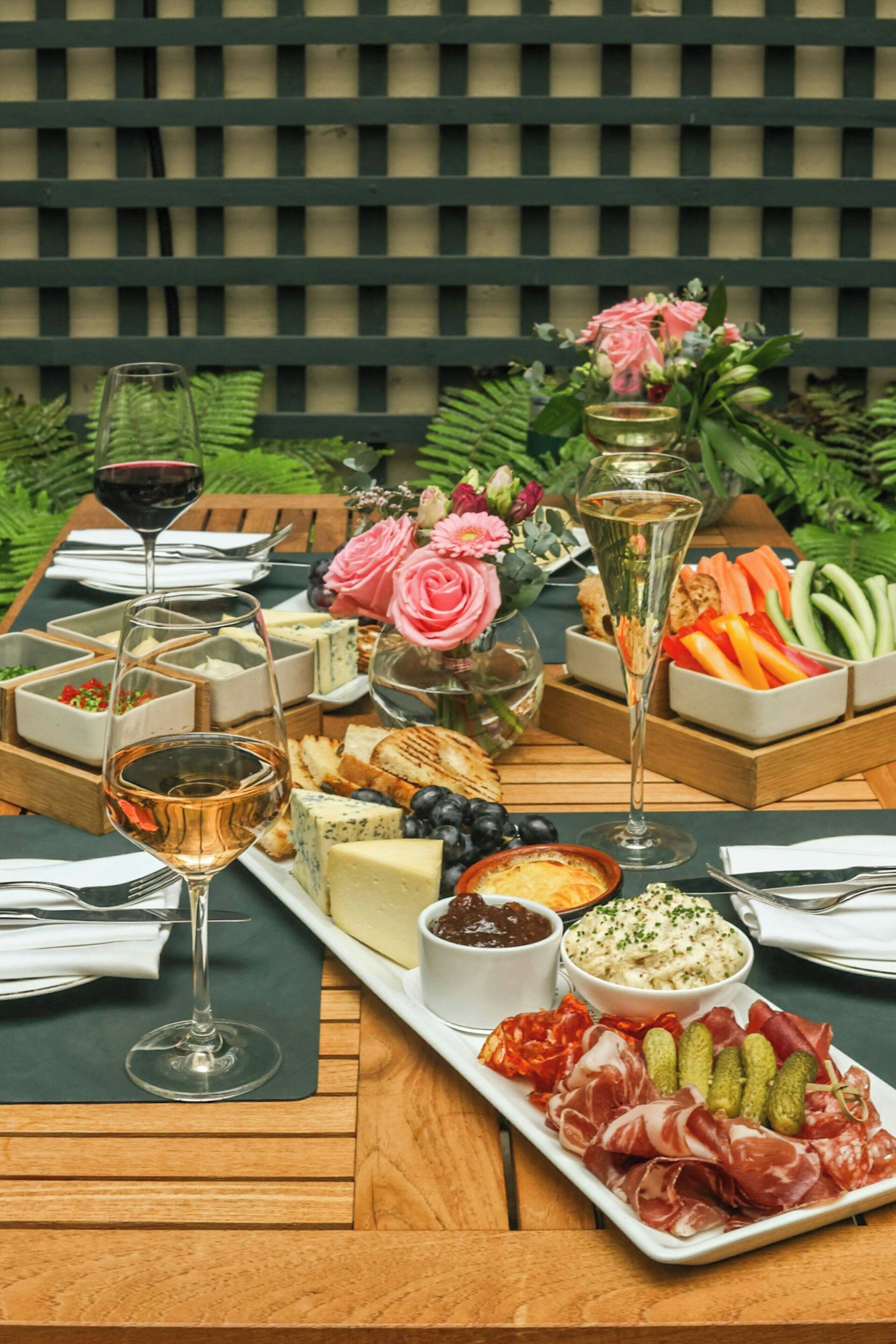Een tafel gedekt met diverse kazen, fruit en vleeswaren, met wijnglazen en een bloemstuk.