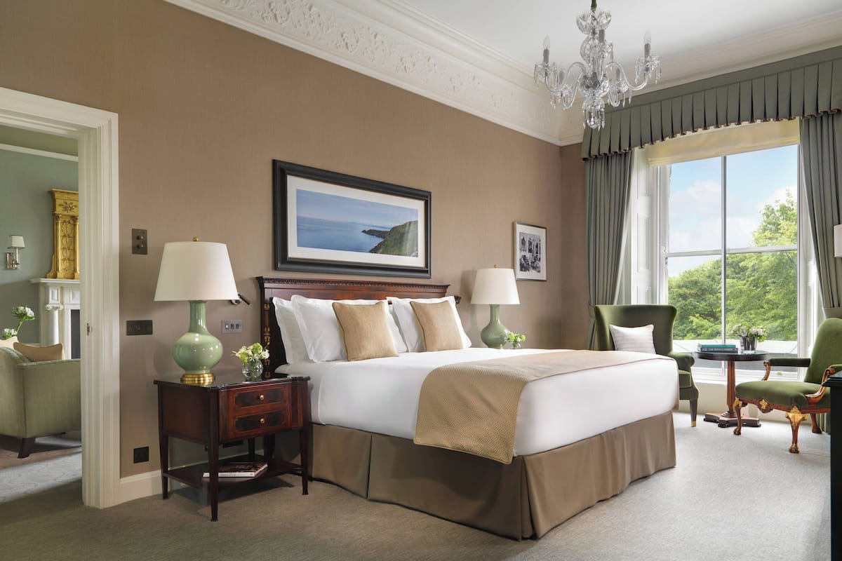 Habitación de hotel con una cama grande, dos mesitas de noche con lámparas, un cuadro en la pared y vistas a zonas verdes por la ventana.