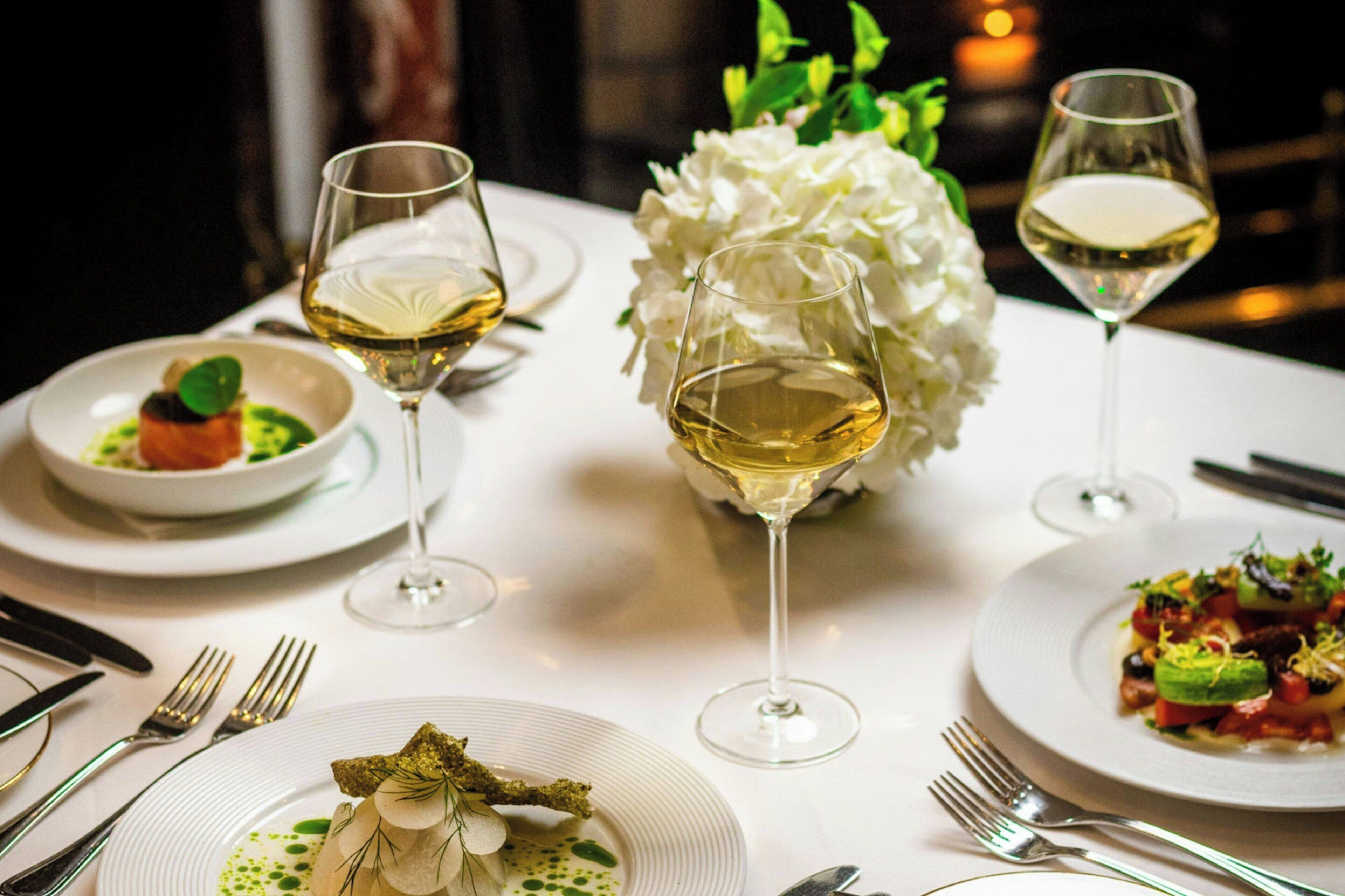 Et spisebord dækket med tallerkener med mad, glas med hvidvin og en blomsterdekoration af hvide blomster.