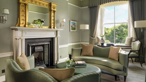 A living room with a fireplace, green sofas, a chandelier, and a large window with curtains.