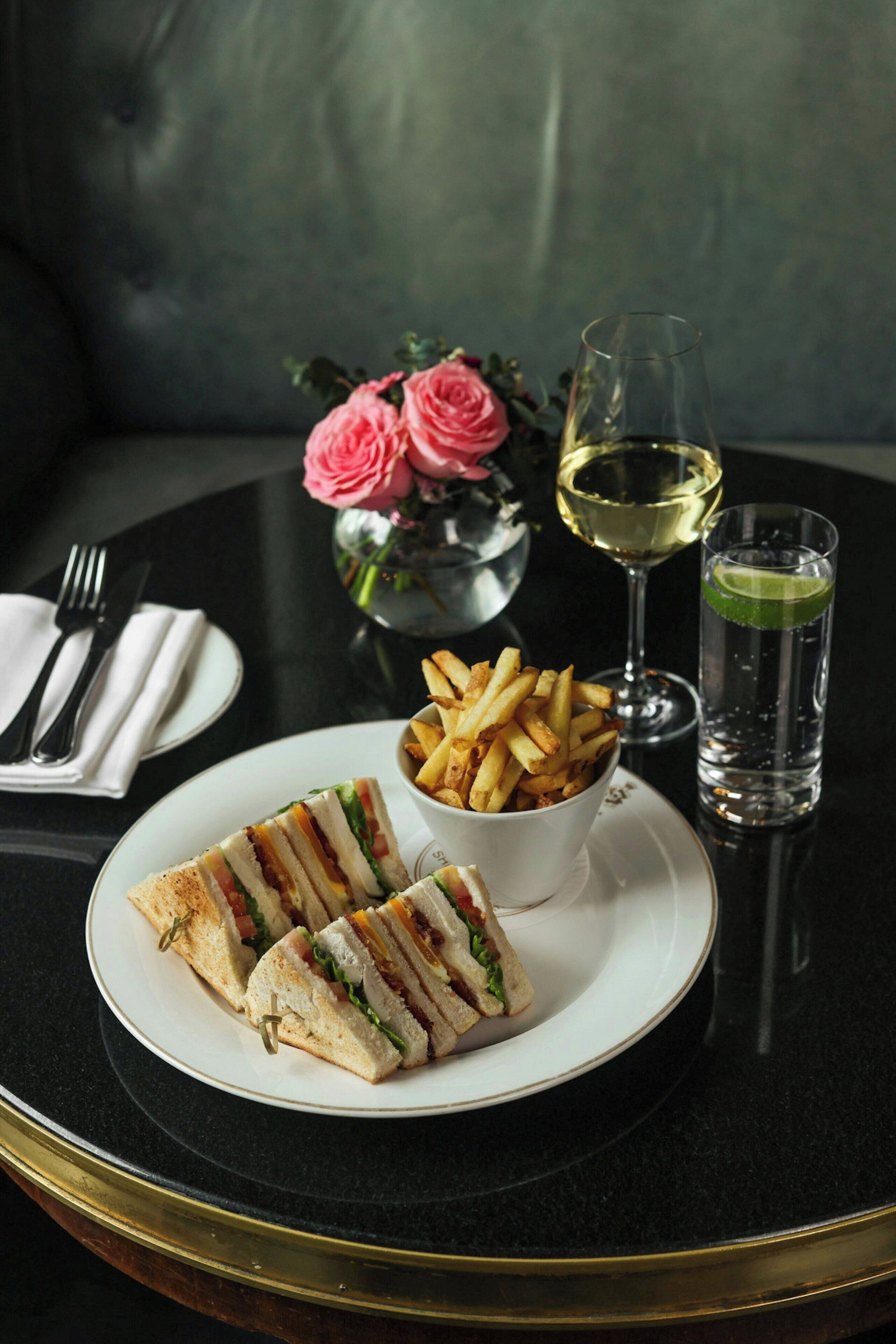 A table with a plate of sandwiches, a bowl of fries, a glass of white wine, and a glass of water.