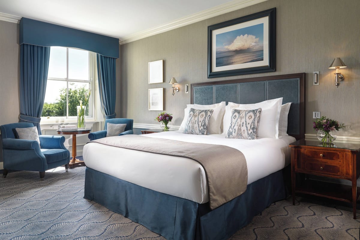 Una habitación de hotel con una cama grande, un sillón azul, una mesita con un florero y una ventana con cortinas.