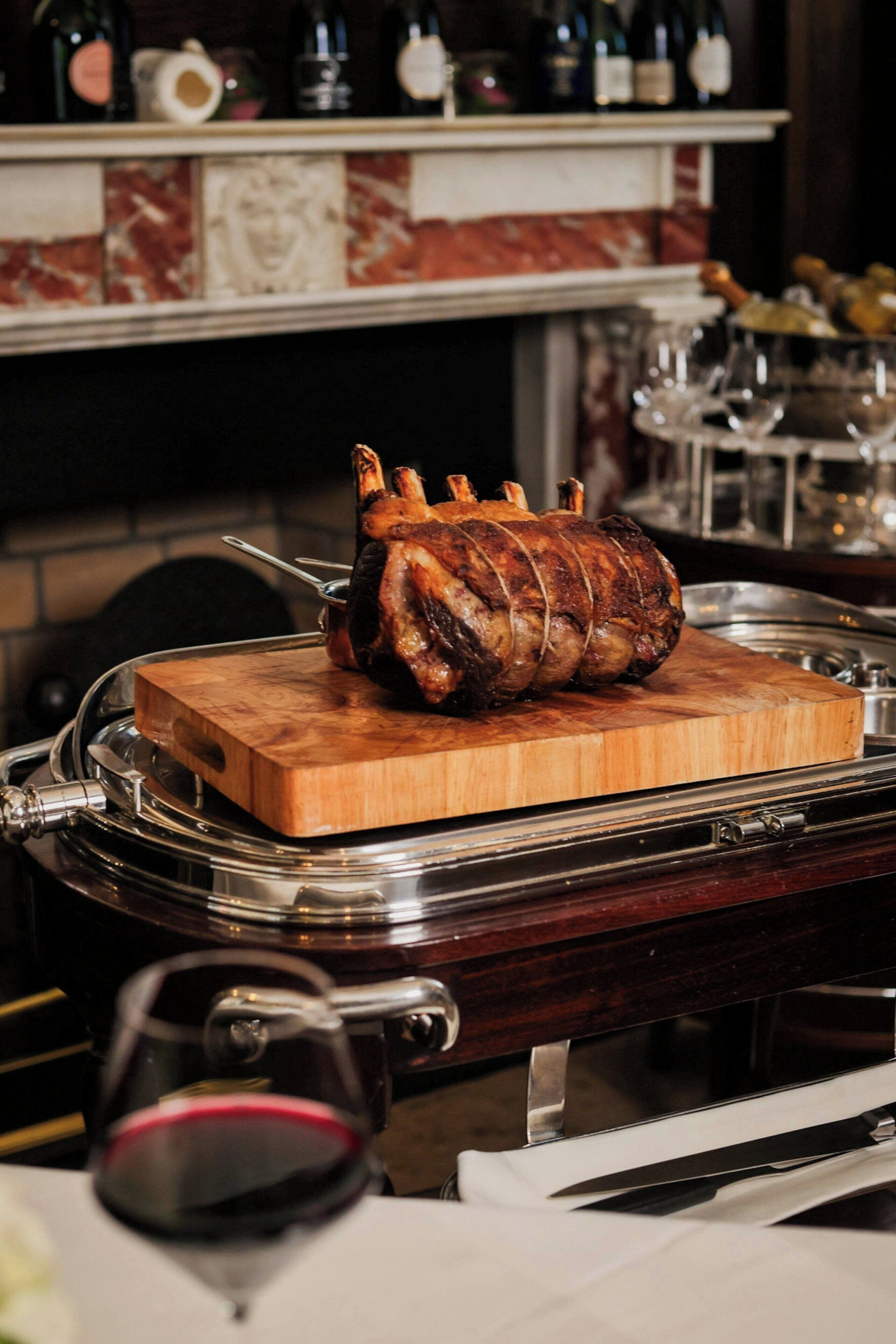 A roasted meat on a wooden board, a wine glass, and a kitchen counter with bottles.