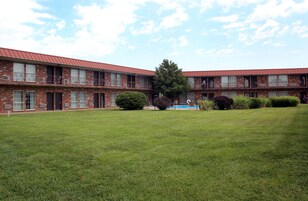 A two-story brick building with a red roof, a courtyard with grass and trees, and a swimming pool in the background.