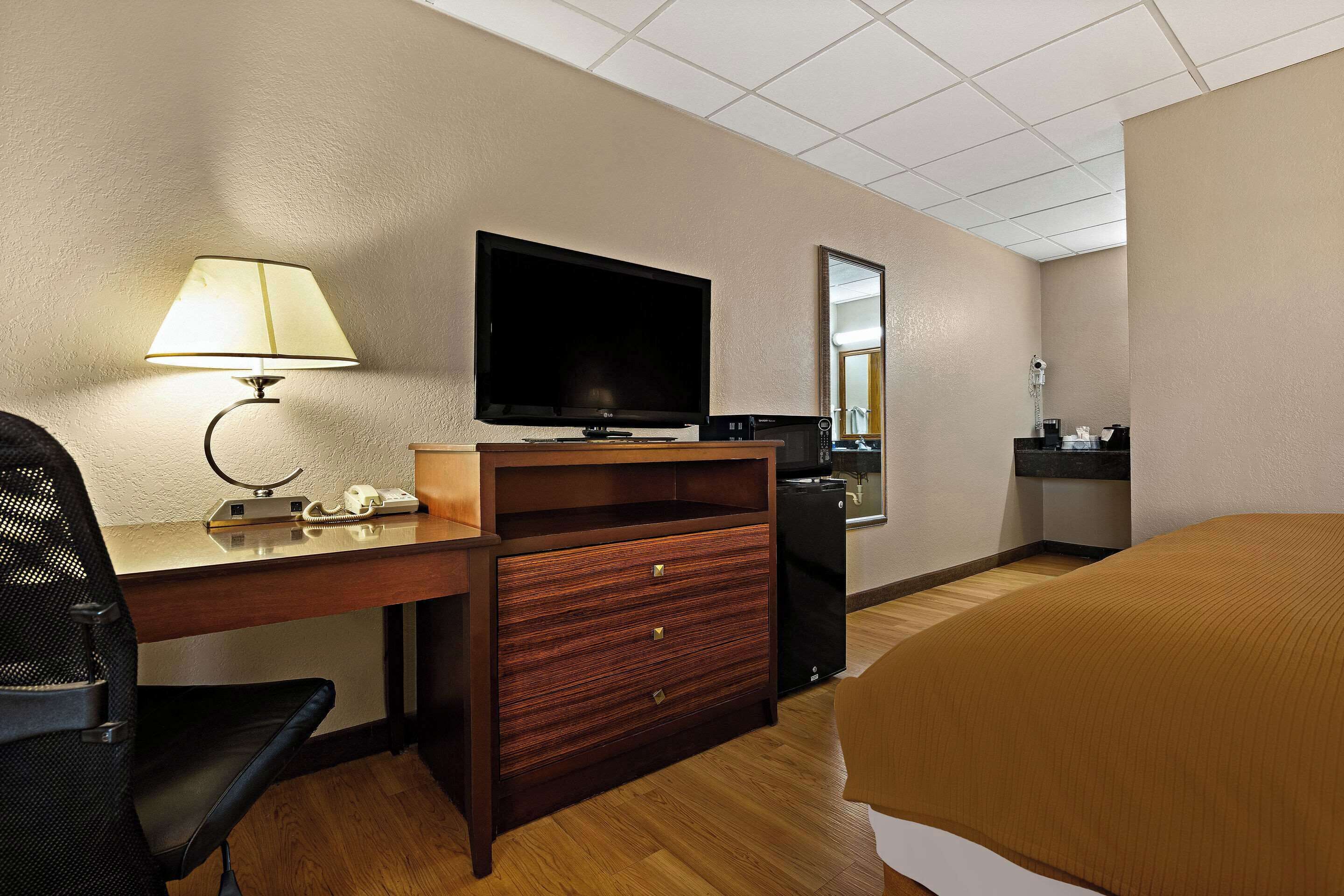 Una habitación de hotel con un escritorio de madera, un televisor de pantalla plana, una lámpara, una cama y una pequeña kitchenette.