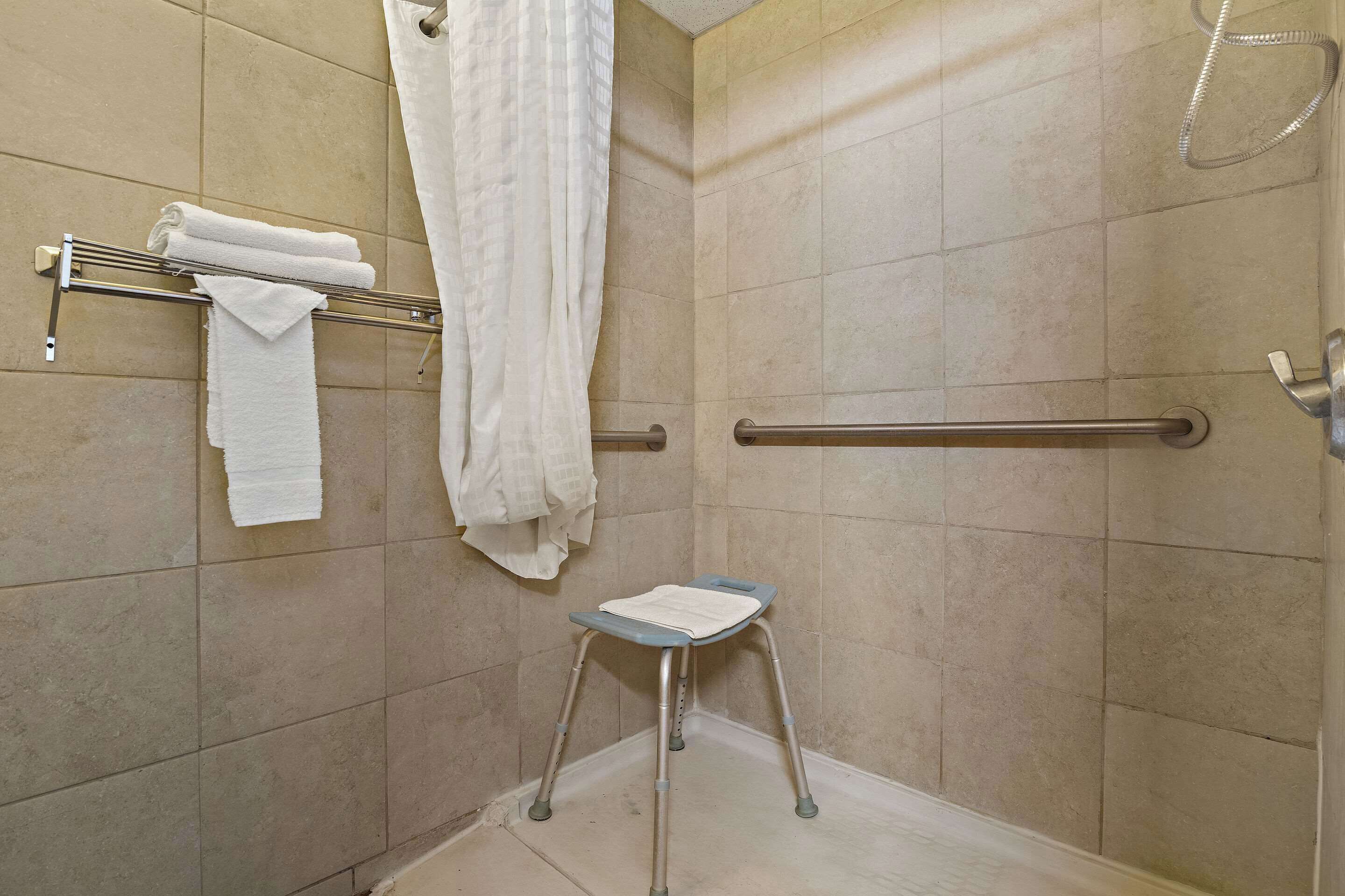 Un baño con azulejos color beige, un toallero blanco, cortina de ducha, barra de apoyo y un taburete pequeño.