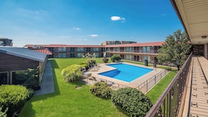 Une cour d’hôtel avec une piscine, de vastes pelouses verdoyantes et un bâtiment en briques surmonté d’un toit rouge.