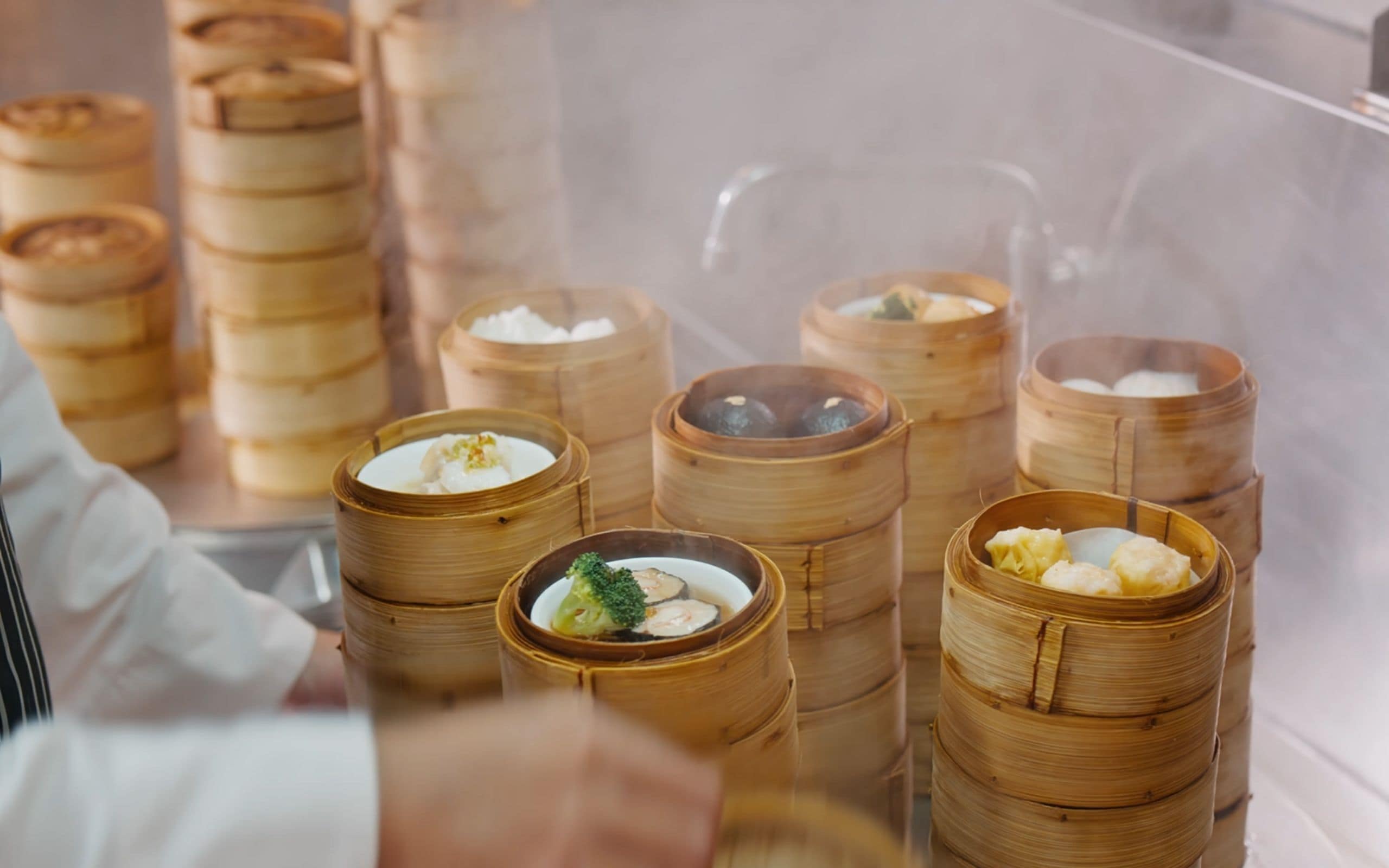 Una hilera de vaporeras de bambú con diversos alimentos visibles a través de una barrera de cristal, probablemente empanadillas o bollos.