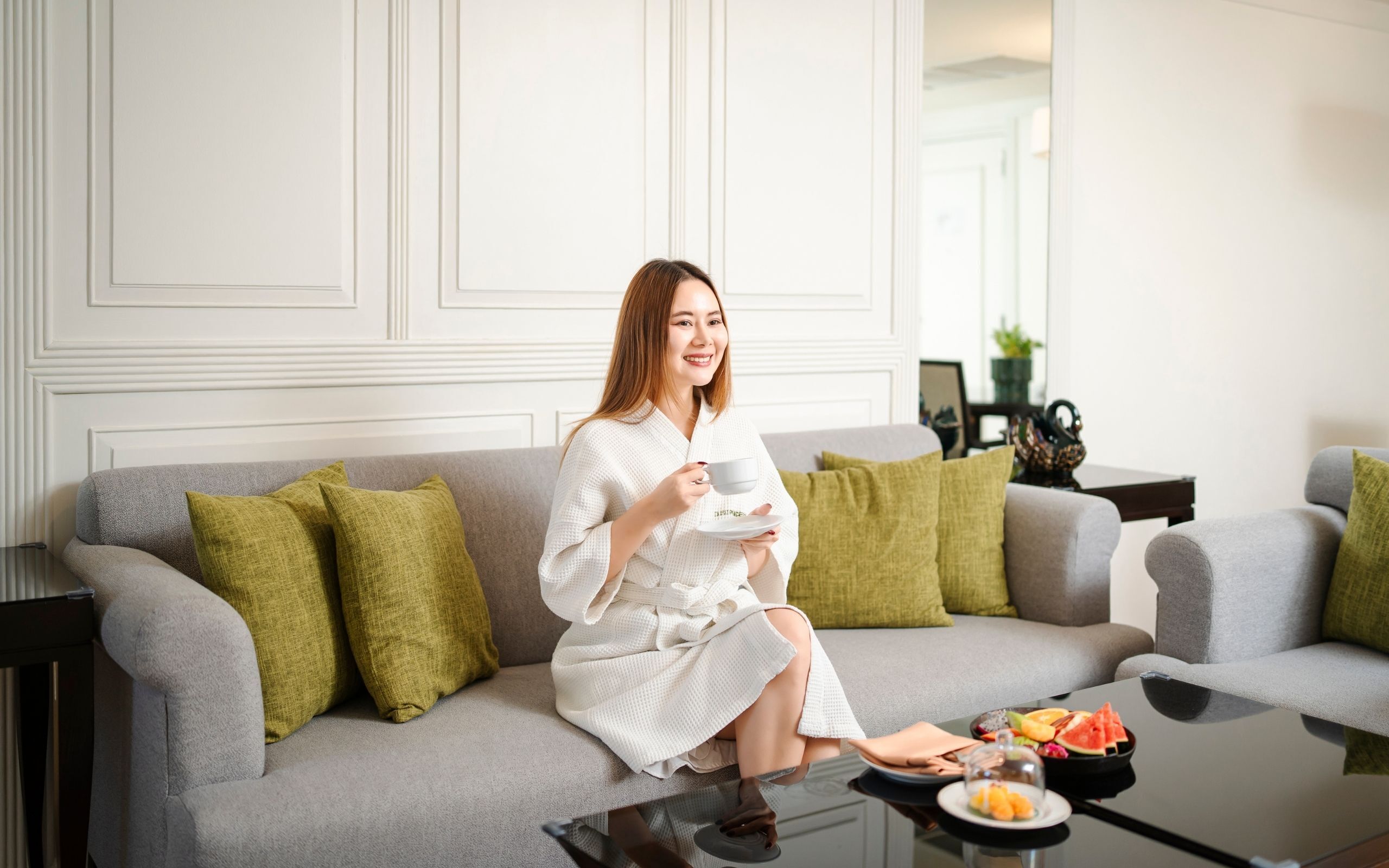 A woman in a white robe sitting on a grey sofa with green cushions, holding a cup and saucer. A table in front of her has a fruit bowl and a glass dome.