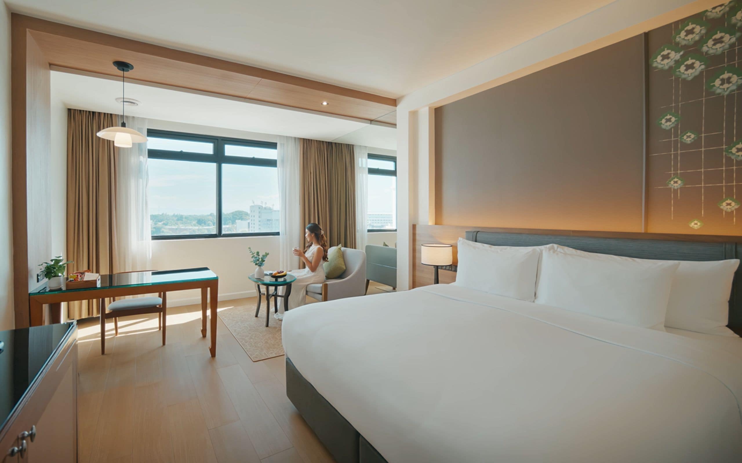 Habitación de hotel con una cama grande, un escritorio, una silla y vistas de la ciudad a través de amplios ventanales.