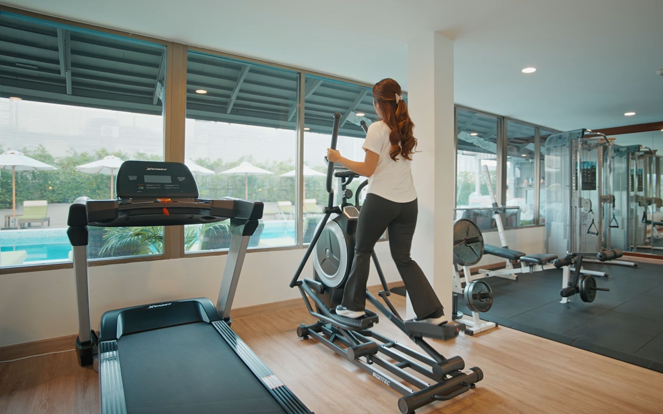 Ein Fitnessraum mit Laufband, Crosstrainer und Krafttrainingsgeräten. Große Fenster bieten Blick auf einen Pool und Grünflächen.