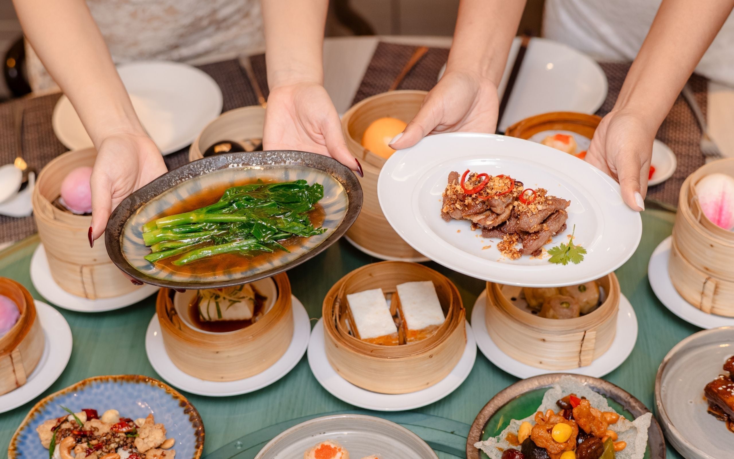 A table with various dishes, including a plate with meat and vegetables, a bowl with greens, and several bamboo steamers.