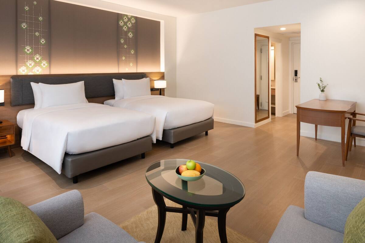 Ein Hotelzimmer mit zwei Betten, einem kleinen Tisch mit Obst, einem Schreibtisch und einem Stuhl.