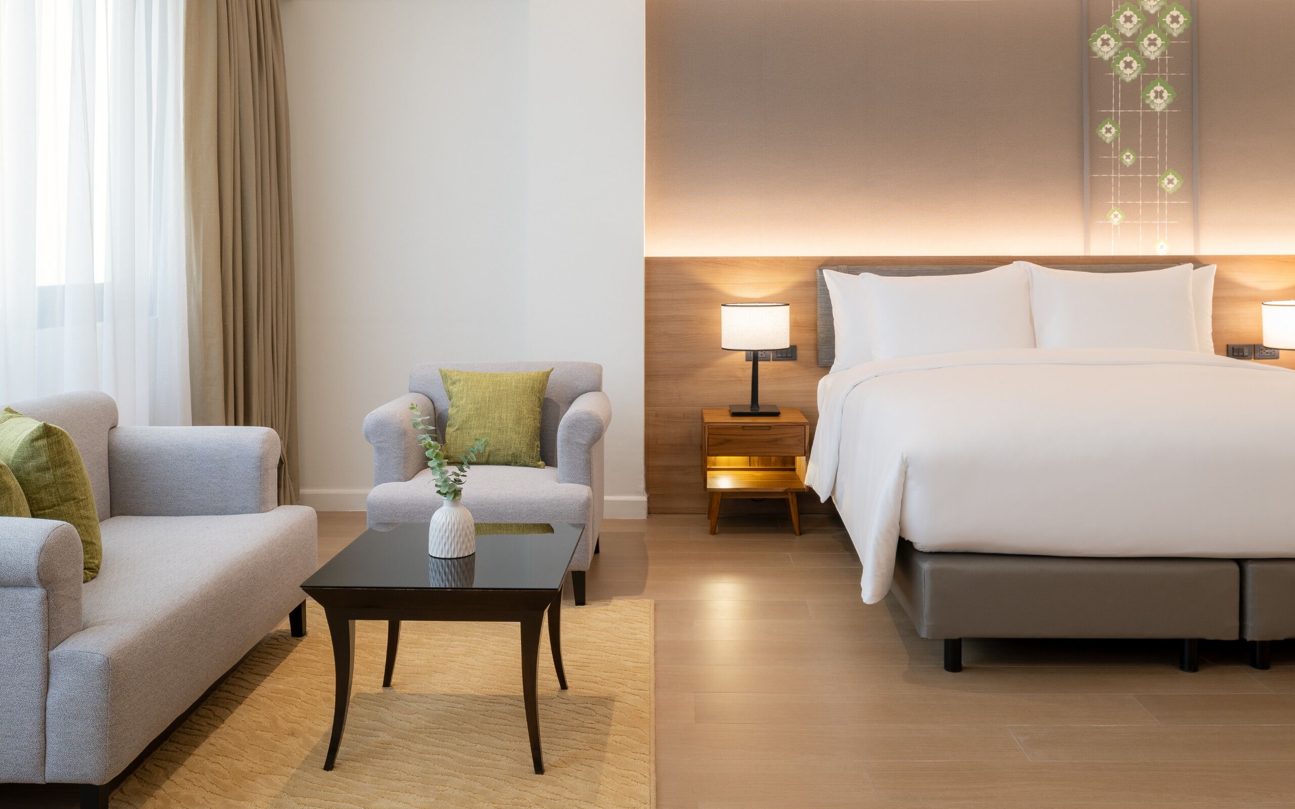 A hotel room with a sofa, armchair, small table, bedside table with lamp, and a bed.