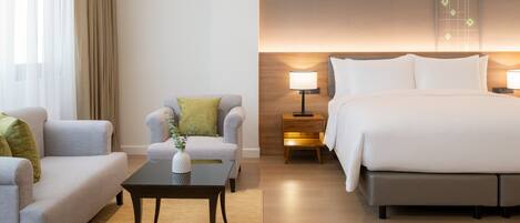 A hotel room with a sofa, armchair, small table, bedside table with lamp, and a bed.