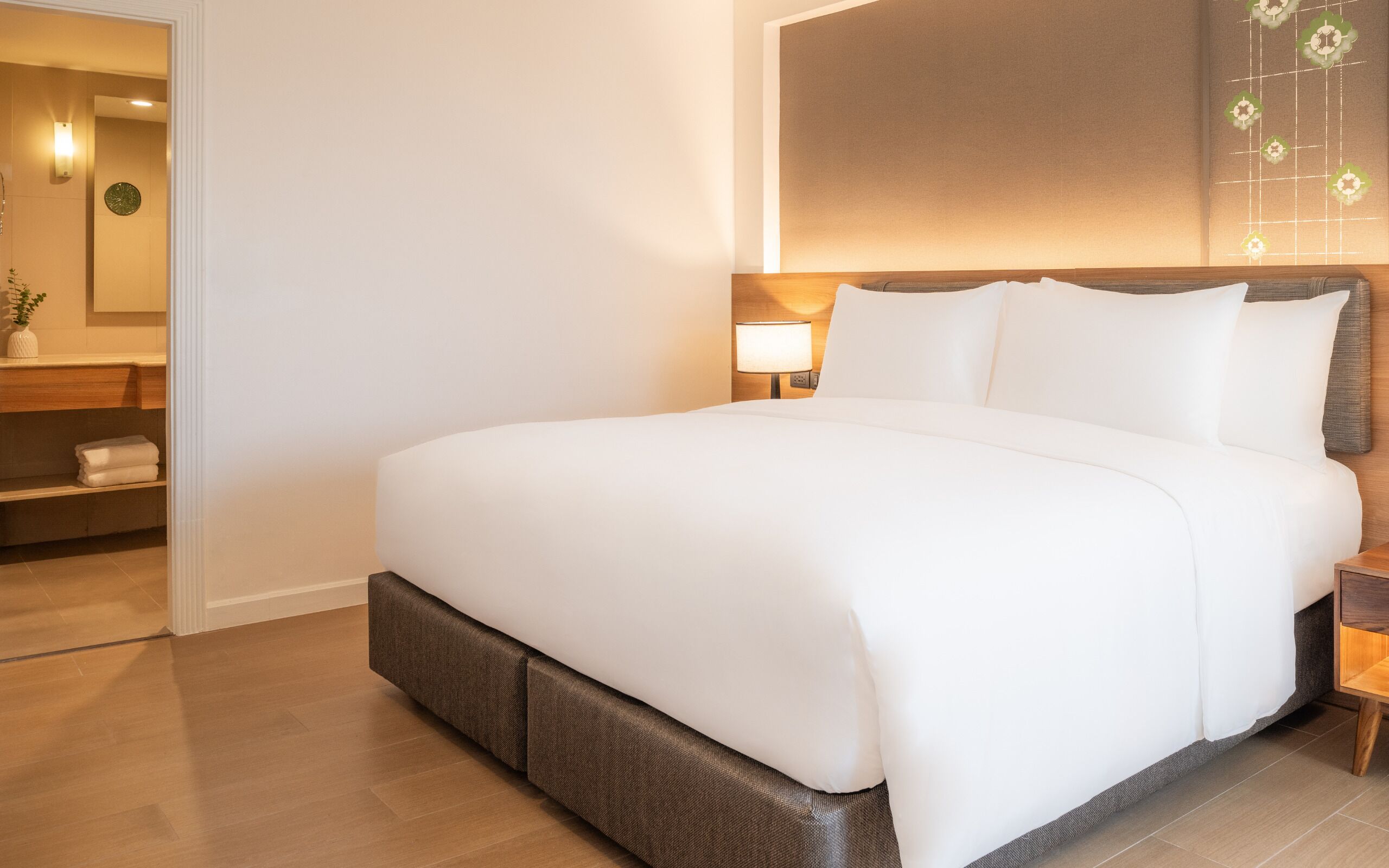 Una cama bien hecha en una habitación de hotel, con cabecera de madera, una mesita de noche con lámpara y un pequeño estante con toallas.