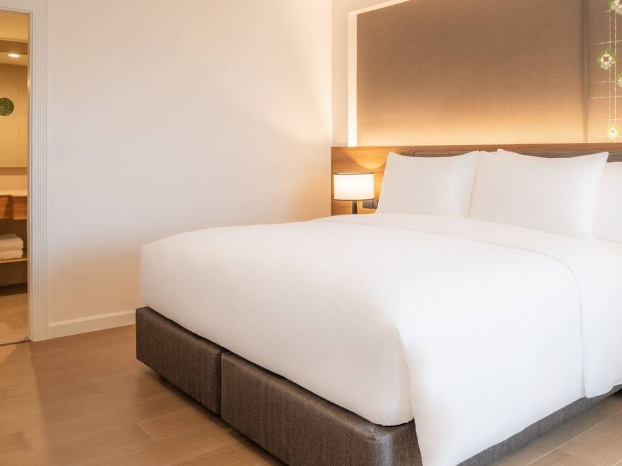 Una cama bien hecha en una habitación de hotel, con cabecera de madera, una mesita de noche con lámpara y un pequeño estante con toallas.