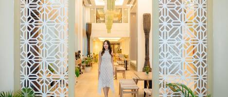 A woman in a white dress standing in a well-lit indoor space with geometric patterned screens, wooden furniture, and hanging light fixtures.