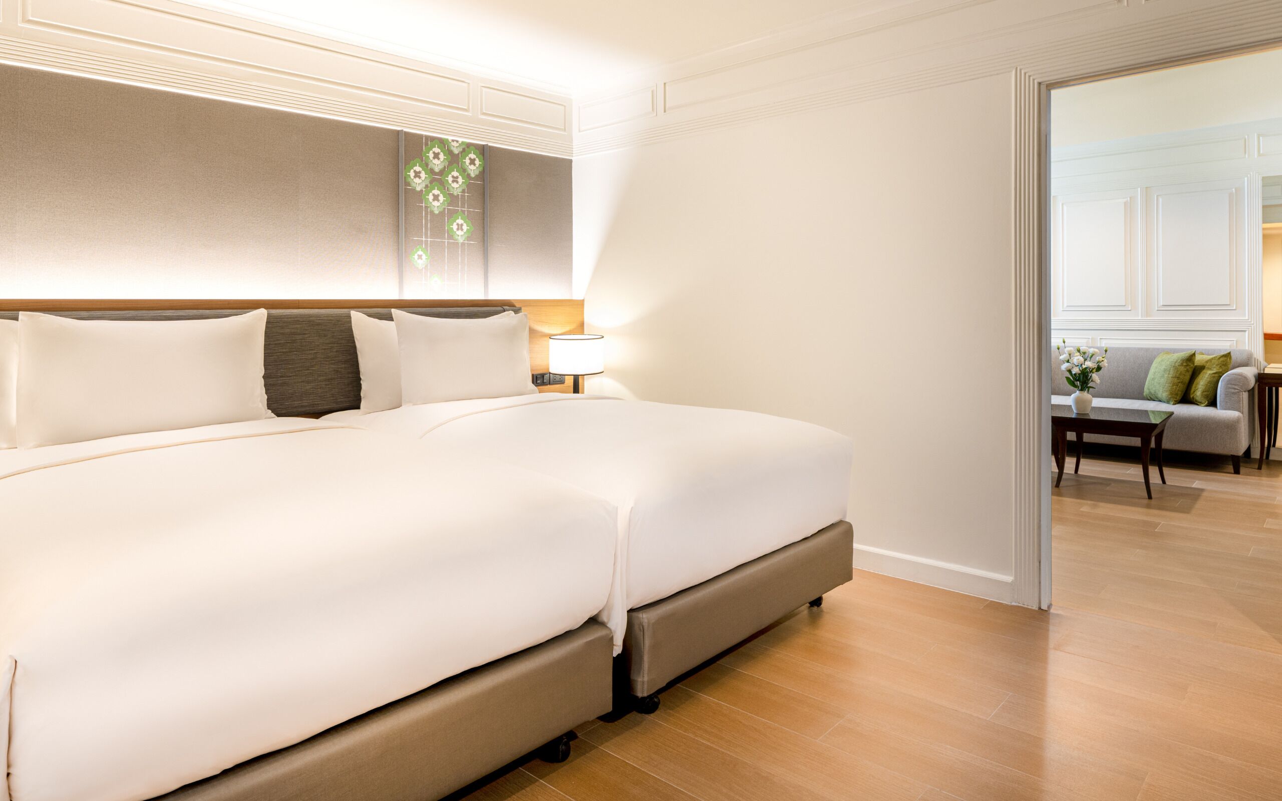 Una cama grande con cabecera, una mesita de noche con lámpara, y vista a un comedor a través de una puerta abierta.