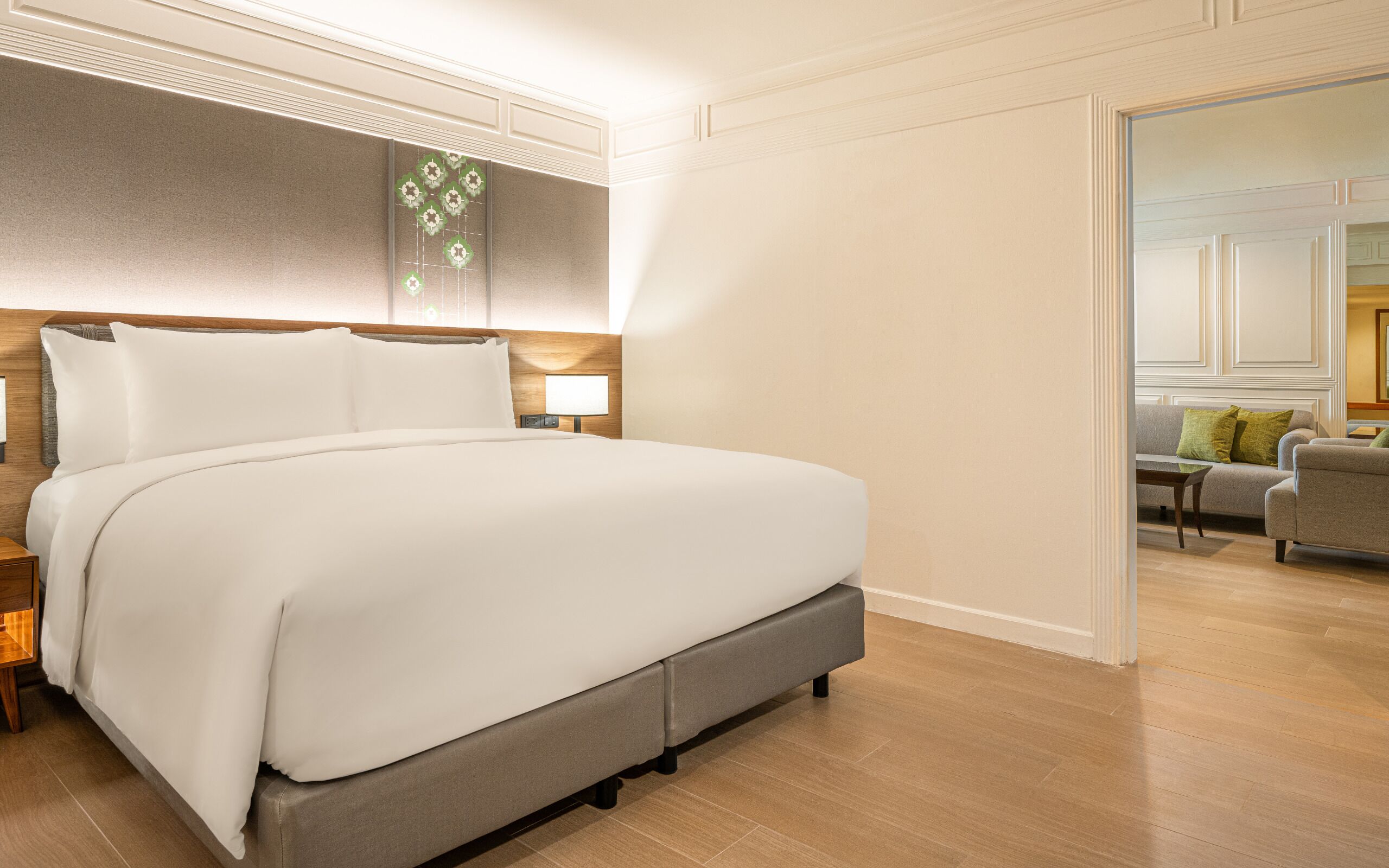A hotel room with a large bed, a nightstand, a lamp, and a view of another room with a sofa and a table.