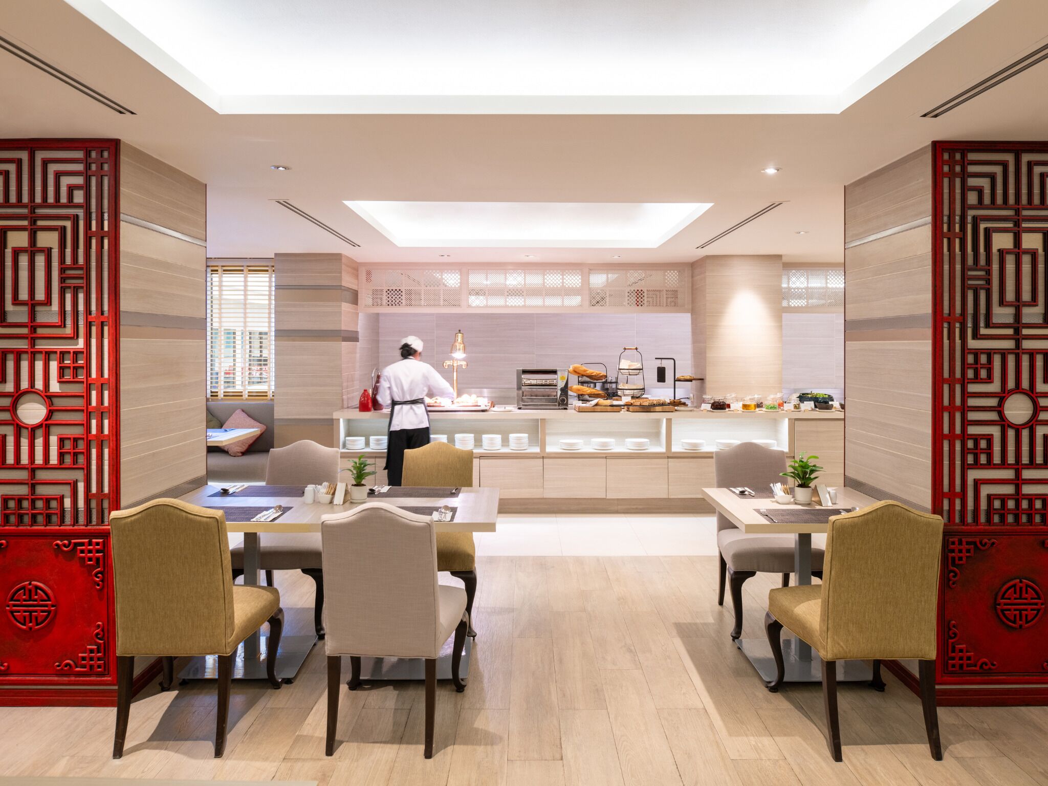 A modern dining area with a chef preparing food at a counter, wooden flooring, and traditional red decor elements.