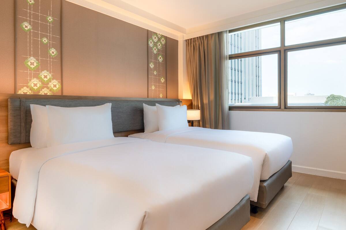 Ein Hotelzimmer mit zwei Betten, einem Nachttisch, einer Lampe, einem Fenster mit Blick auf Gebäude und einer dekorativen Wandverkleidung.