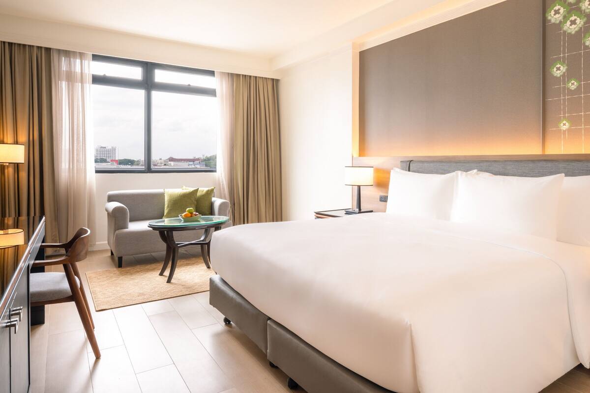 Ein Hotelzimmer mit einem großen Bett, einem Schreibtisch mit Stuhl, einem kleinen Tisch mit Obstschale und Blick auf die Stadt durch das Fenster.
