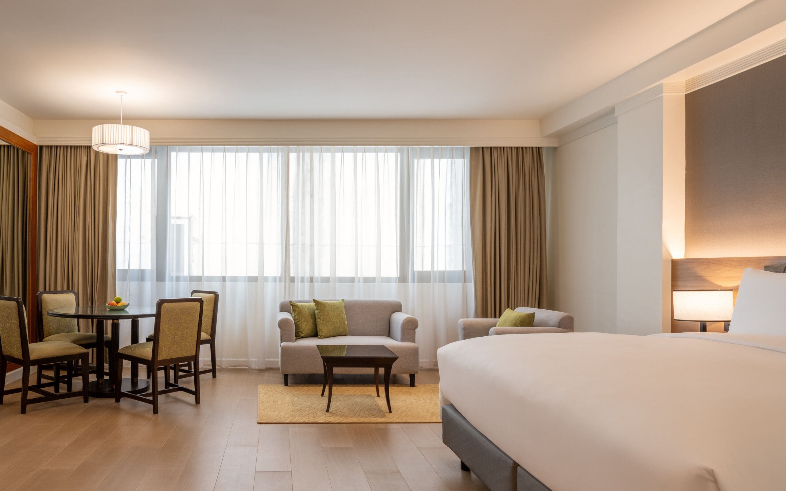 A hotel room with a large bed, two armchairs, a small table, and a sofa. There is a window with sheer curtains and a view of the outside.