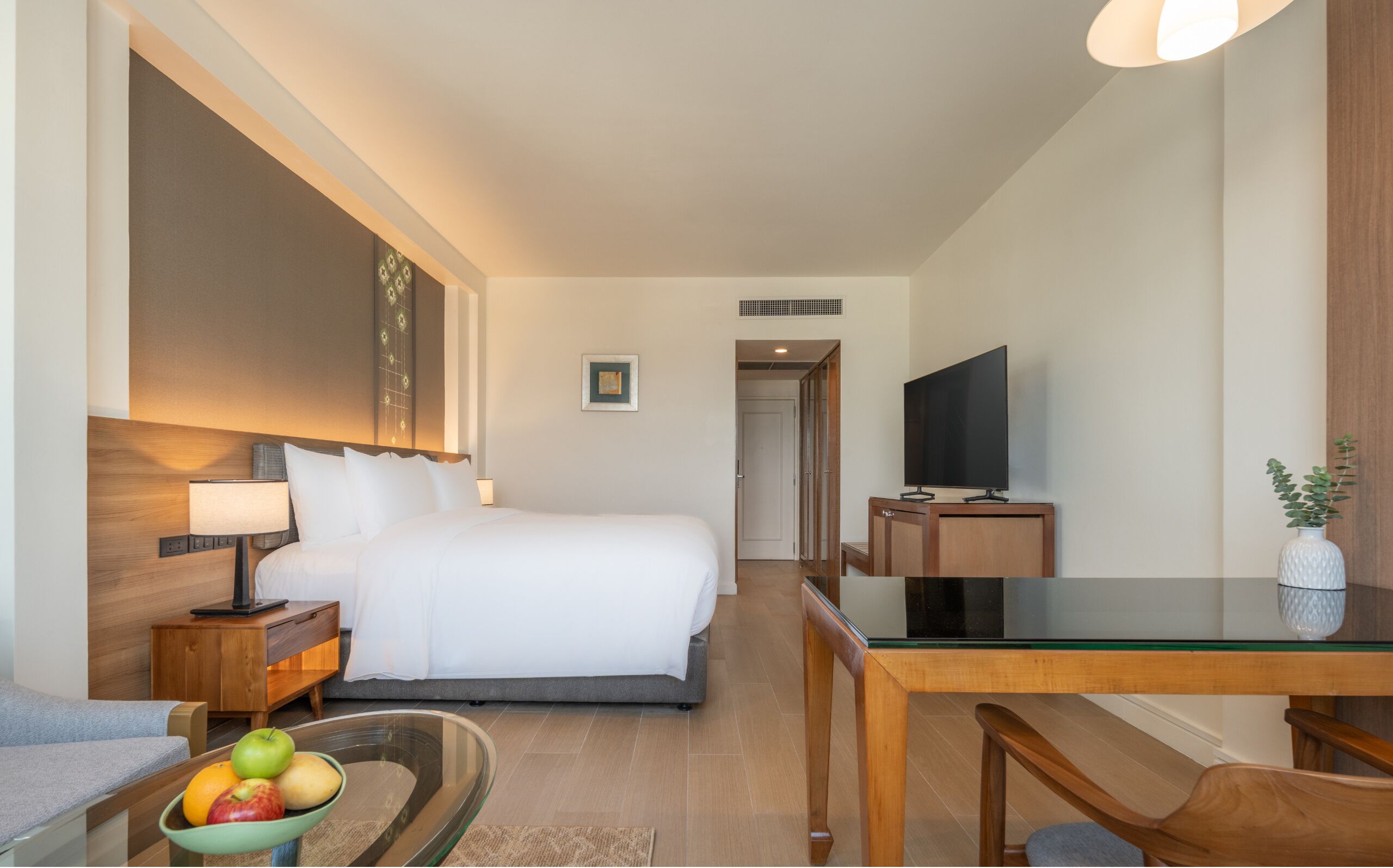 A hotel room with a bed, bedside table, lamp, TV, dining table, and a bowl of fruit.