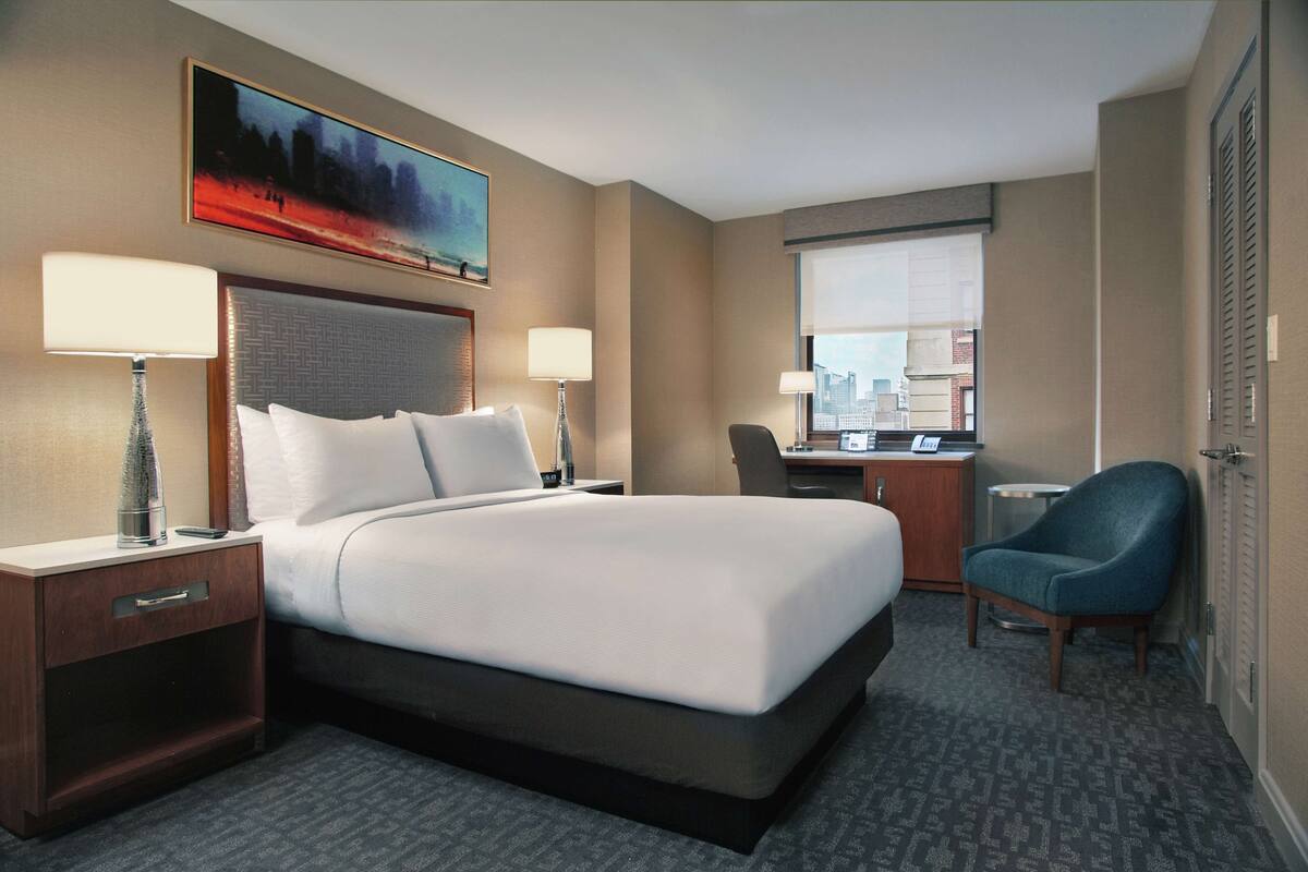 Una habitación de hotel con una cama grande, dos mesitas de noche con lámparas, un escritorio con silla y una ventana con vistas a la ciudad.
