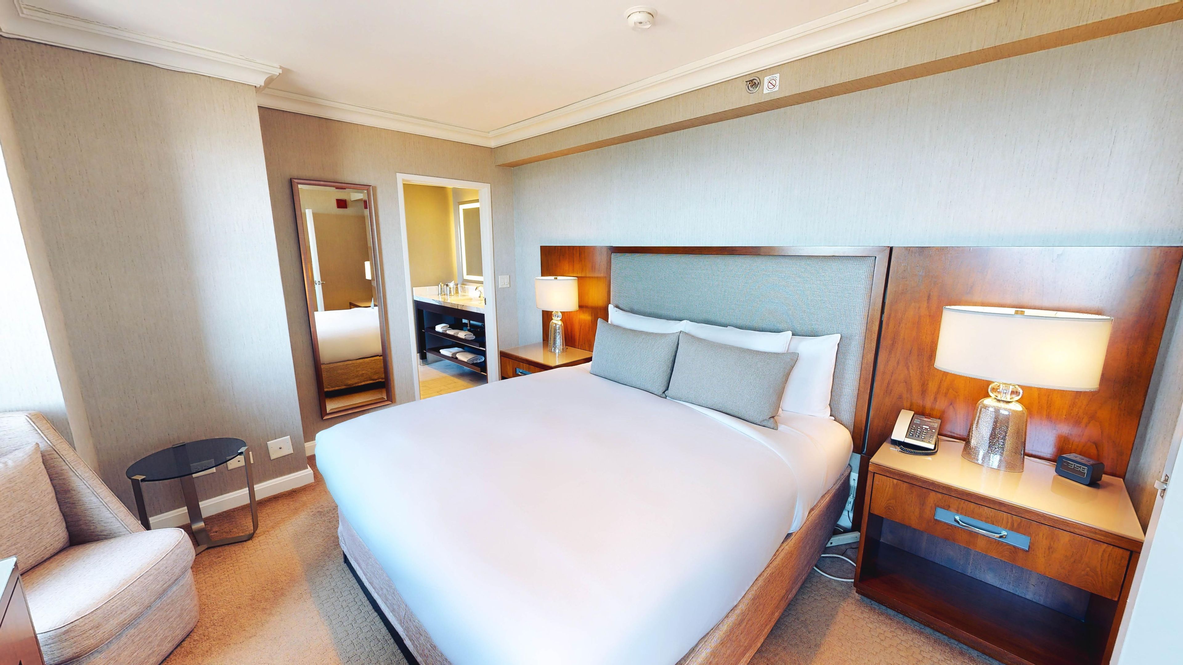 Een hotelkamer met een groot bed, een nachtkastje met een lamp, een telefoon op een nachtkastje, een spiegel en een stoel.